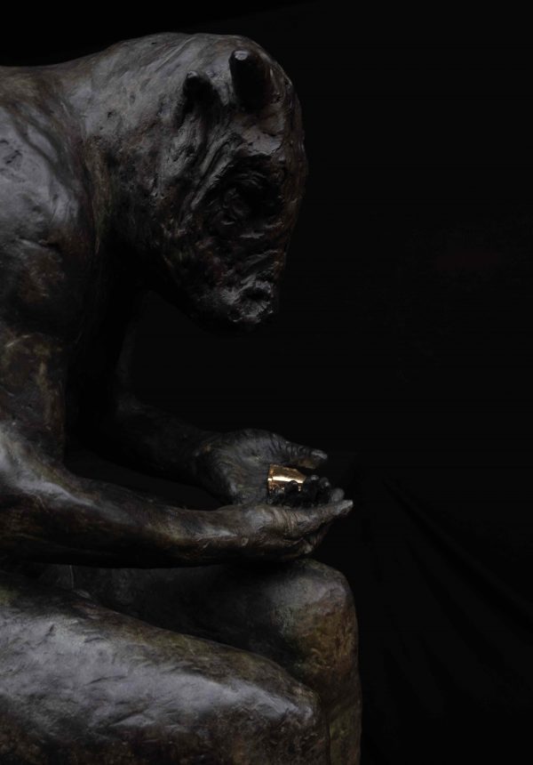 Bronze sculpture of a seated minotaur bent over, reading intently from a small and gold-colored book he holds in his hands, by Beth Carter titled Reading Minotaur IV.